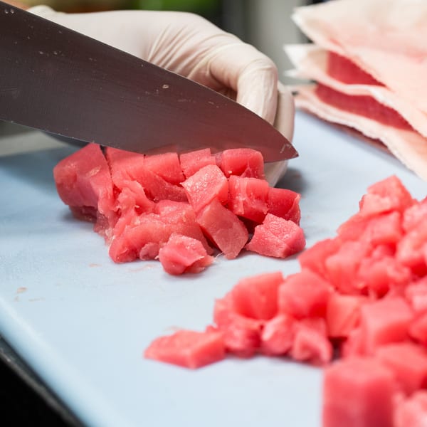 Gloved hands dicing fresh red tuna on a light blue cutting board for Grindz poke.