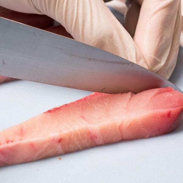 Gloved hands slicing a fresh piece of raw tuna on a white cutting board for poke.